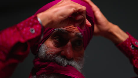 Primer-Plano,-Toma-De-Estudio-Con-Iluminación-Tenue-De-Un-Hombre-Sikh-Mayor-Con-Barba-Atando-Una-Tela-Para-Un-Turbante-Contra-Un-Fondo-Oscuro,-Filmado-En-Tiempo-Real